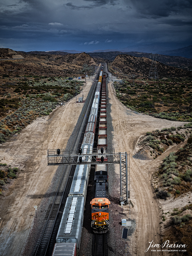 September 20th, 2024, A BNSF freight and a BNSF rock train pass each other at the Summit of Cajon Pass in southern California and the BNSF Cajon Subdivision.

According to Wikipedia: Cajon Pass is a mountain pass between the San Bernardino Mountains to the east and the San Gabriel Mountains to the west in Southern California. Created by the movements of the San Andreas Fault, it has an elevation of 3,777 ft (1,151 m). Located in the Mojave Desert, the pass is an important link from the Greater San Bernardino Area to the Victor Valley, and northeast to Las Vegas. The Cajon Pass area is on the Pacific Crest Trail.

Cajon Pass is at the head of Horsethief Canyon, traversed by California State Route 138 (SR 138) and railroad tracks owned by BNSF Railway and Union Pacific Railroad. Improvements in 1972 reduced the railroad's maximum elevation from about 3,829 to 3,777 feet while reducing curvature. Interstate 15 does not traverse Cajon Pass, but rather the nearby Cajon Summit. The entire area, Cajon Pass and Cajon Summit, is often referred to as Cajon Pass, but a distinction is made between Cajon Pass and Cajon Summit.

The California Southern Railroad, a subsidiary of the Atchison, Topeka and Santa Fe Railway, was the first railroad through Cajon Pass. The line through the pass was built in the early 1880s to connect the present-day cities of Barstow and San Diego. Today the Union Pacific Railroad and BNSF Railway (the successor to the Santa Fe) use the pass to reach Los Angeles and San Bernardino as part of the Southern Transcon. Due to the many trains, scenery and easy access, it is a popular location for railfans, and many photographs of trains on Cajon Pass appear in books and magazines.

Tech Info: DJI Mavic 3 Classic Drone, RAW, 24mm, f/2.8, 1/1600, ISO 100.

#railroad #railroads #train, #trains #railway #railway #steamtrains #railtransport #railroadengines #picturesoftrains #picturesofrailways #besttrainphotograph #bestphoto #photographyoftrains #bestsoldpicture #JimPearsonPhotography #trainsfromtheair #trainsfromadrone #CajonPass