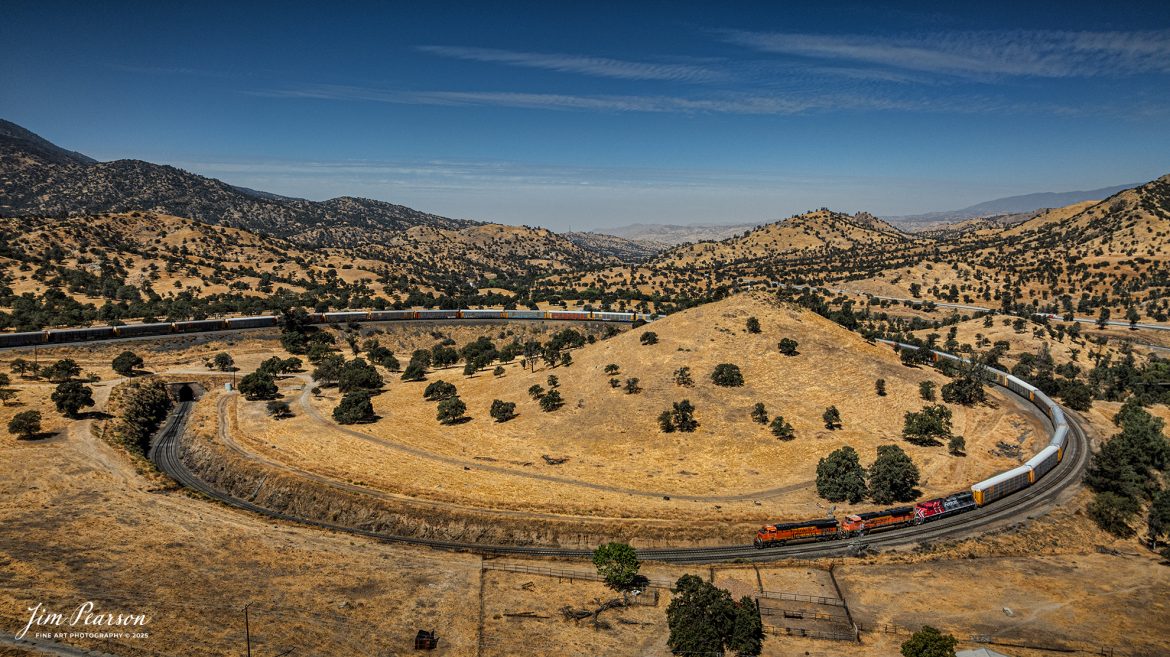 September 18th, 2024, BNSF 7115, 9128 and Ferromex 4050 lead an northbound train through Tehachapi Loop on the Union Pacific Mojave Subdivision at Keene, California.

According to Wikipedia: The Tehachapi Loop is a 3,779-foot-long (0.72 mi; 1.15 km) spiral, or helix, on the Union Pacific Railroad Mojave Subdivision through Tehachapi Pass, of the Tehachapi Mountains in Kern County, south-central California. The line connects Bakersfield and the San Joaquin Valley to Mojave in the Mojave Desert.

Rising at a steady two-percent grade, the track gains 77 feet (23 m) in elevation and makes a 1,210-foot-diameter (370 m) circle. Any train that is more than 3,800 feet (1,200 m) long—about 56 boxcars—passes over itself going around the loop. At the bottom of the loop, the track passes through Tunnel 9, the ninth tunnel built as the railroad was extended from Bakersfield.

The line averages about 36 freight trains each day. Passenger trains such as Amtrak's San Joaquin are banned from the loop, although the Coast Starlight can use it as a detour. Its frequent trains and scenic setting make the Tehachapi Loop popular with railfans. In 1998, it was named a National Historic Civil Engineering Landmark. It is also designated as California Historical Landmark #508.

One of the engineering feats of its day, the Loop was built by Southern Pacific Railroad to ease the grade over Tehachapi Pass. Construction began in 1874, and the line opened in 1876.

Tech Info: DJI Mavic 3 Classic Drone, RAW, 24mm, f/2.8, 1/2500, ISO 100.

#railroad #railroads #train, #trains #railway #railway #steamtrains #railtransport #railroadengines #picturesoftrains #picturesofrailways #besttrainphotograph #bestphoto #photographyoftrains #bestsoldpicture #JimPearsonPhotography #trainsfromtheair #trainsfromadrone #TehachapiLoop