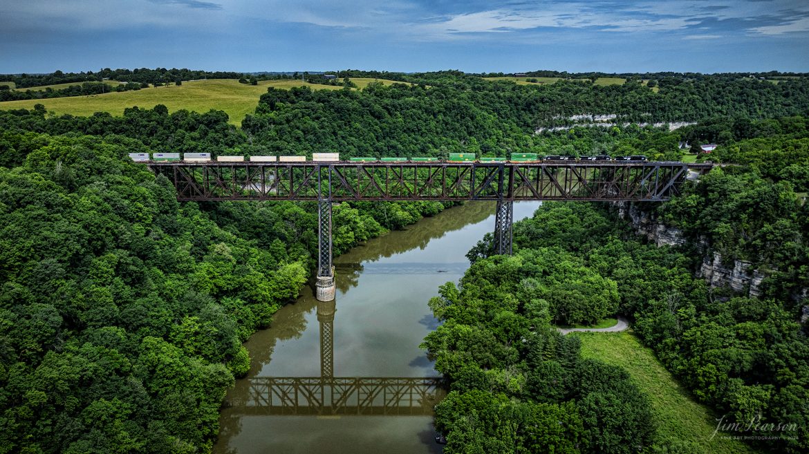 Norfolk Southern intermodal 28C heads northbound across High Bridge on June 8th, 2024, crossing the Kentucky River on the NS CNO&TP First District at Highbridge, Kentucky.

According to Wikipedia: The High Bridge is a railroad bridge crossing the Kentucky River Palisades, that rises approximately 275 feet from the river below and connects Jessamine and Mercer counties in Kentucky. Formally dedicated in 1879, it is the first cantilever bridge constructed in the United States. It has a three-span continuous under-deck truss used by Norfolk Southern Railway to carry trains between Lexington and Danville. It has been designated as a National Historic Civil Engineering Landmark.

In 1851, the Lexington & Danville Railroad, with Julius Adams as chief engineer, retained John A. Roebling to build a railroad suspension bridge across the Kentucky River for a line connecting Lexington and Danville, Kentucky west of the intersection of the Dix and Kentucky rivers. In 1855, the company ran out of money and the project was resumed by Cincinnati Southern Railroad in 1873 following a proposal by C. Shaler Smith for a cantilever design using stone towers designed by John A. Roebling (who designed the Brooklyn Bridge).

The bridge was erected using a cantilever design with a three-span continuous under-deck truss and was opened in 1877 on the Cincinnati Southern Railway. It was 275 feet (84 m) tall and 1,125 feet (343 m) long: the tallest bridge above a navigable waterway in North America and the tallest railroad bridge in the world until the early 20th century. Construction was completed using 3,654,280 pounds of iron at a total cost of $404,373.31. In 1879 President Rutherford B. Hayes and Gen. William Tecumseh Sherman attended the dedication.

After years of heavy railroad use, the bridge was rebuilt by Gustav Lindenthal in 1911. Lindenthal reinforced the foundations and rebuilt the bridge around the original structure. To keep railroad traffic flowing, the track deck was raised by 30 feet during construction and a temporary trestle was constructed. In 1929, an additional set of tracks was built to accommodate increased railroad traffic and the original limestone towers were removed.

Tech Info: DJI Mavic 3 Classic Drone, RAW, 24mm, f/2.8, 1/2000, ISO 160.

#trainphotography #railroadphotography #trains #railways #trainphotographer #railroadphotographer #jimpearsonphotography #NorfolkSouthern #KentuckyTrains