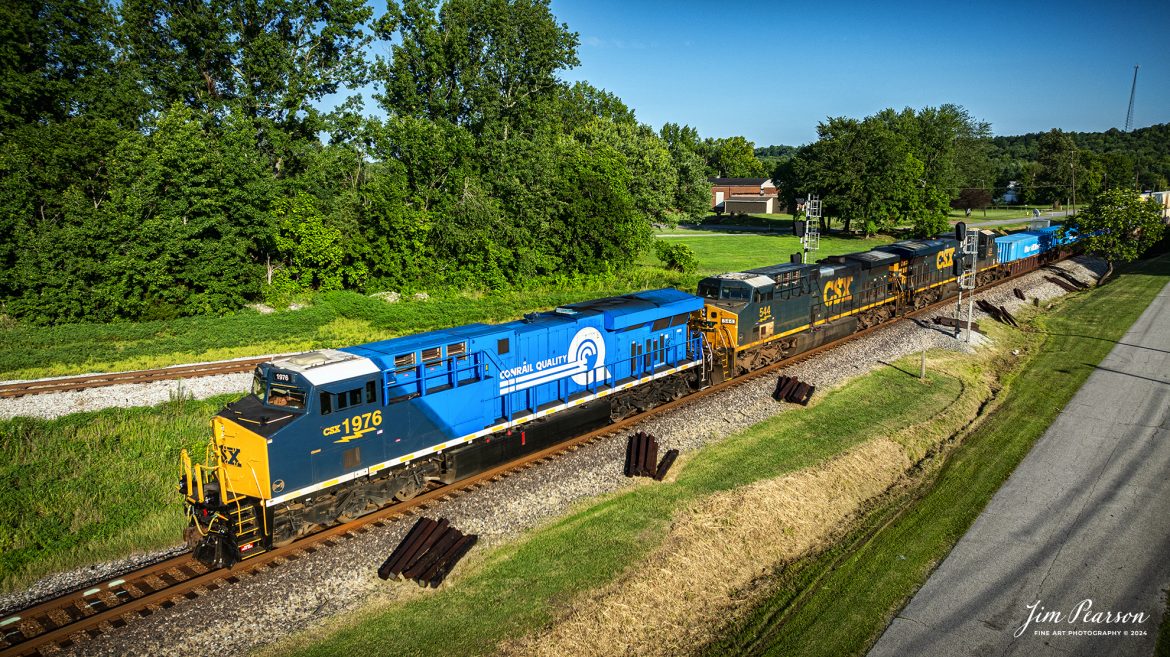 The CSX Conrail Heritage Locomotive leads CSX I026 northbound through Mortons Junction at Mortons Gap, Ky, on July 18th, 2024, on the CSX Henderson Subdivision as they make their daily run to Chicago, Illinois. Next to the engine house is RJC Genset 2698.

According to a CSX Press Release: July 22, 2023 – CSX has unveiled the fourth locomotive in its series of custom paint designs that honor its heritage railroads. The latest in the series commemorates the Consolidated Rail Corporation, or Conrail, and bears the number 1976, the year the railroad was founded.

CSX and Norfolk Southern acquired and divided most of the Conrail network in 1997. Today, the company continues to exist as a terminal and switching railroad for CSX and NS customers in North Jersey-New York City metro area.

The Conrail paint scheme of a double-lined C inside a large, bold Q was a fixture for many years across the Northeast where the majority of the railroad’s operations were centered. The heritage locomotive design conceived and executed at CSX’s locomotive shop in Waycross, Georgia, recreates the iconic “Conrail Quality” logo and features Conrail blue on the rear two-thirds of the engine, then transitions to the current CSX colors and design on the cab portion of the unit.

CSX 1976 and other locomotives in the heritage series will be placed into revenue service and travel CSX’s 20,000 rail network, reinforcing employee pride in the history of the railroad that continues to move the nation’s economy with safe, reliable and sustainable rail-based transportation services.

Tech Info: DJI Mavic 3 Classic Drone, RAW, 22mm, f/2.8, 1/1250, ISO 100.

#railroad #railroads #train #trains #bestphoto #railroadengines #picturesoftrains #picturesofrailway #bestphotograph #photographyoftrains #trainphotography #JimPearsonPhotography #trendingphoto