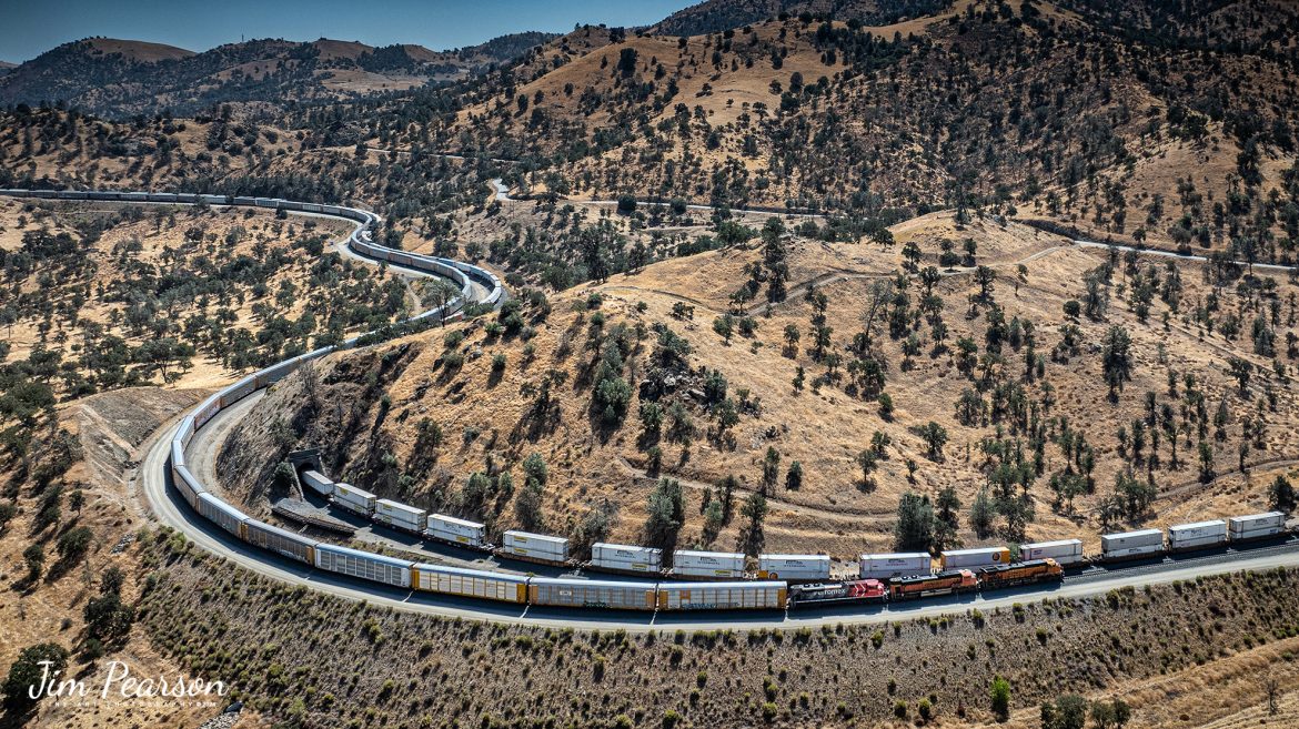 September 18th, 2024, BNSF 7115, 9128 and Ferromex 4050 lead an westbound train next to tunnel 10, as they wait for an eastbound intermodal to clear Tehachapi Loop on the Union Pacific Mojave Subdivision at Keene, California.

According to Wikipedia: The Tehachapi Loop is a 3,779-foot-long (0.72 mi; 1.15 km) spiral, or helix, on the Union Pacific Railroad Mojave Subdivision through Tehachapi Pass, of the Tehachapi Mountains in Kern County, south-central California. The line connects Bakersfield and the San Joaquin Valley to Mojave in the Mojave Desert.

Rising at a steady two-percent grade, the track gains 77 feet (23 m) in elevation and makes a 1,210-foot-diameter (370 m) circle. Any train that is more than 3,800 feet (1,200 m) long—about 56 boxcars—passes over itself going around the loop. At the bottom of the loop, the track passes through Tunnel 9, the ninth tunnel built as the railroad was extended from Bakersfield.

The line averages about 36 freight trains each day. Passenger trains such as Amtrak's San Joaquin are banned from the loop, although the Coast Starlight can use it as a detour. Its frequent trains and scenic setting make the Tehachapi Loop popular with railfans. In 1998, it was named a National Historic Civil Engineering Landmark. It is also designated as California Historical Landmark #508.

One of the engineering feats of its day, the Loop was built by Southern Pacific Railroad to ease the grade over Tehachapi Pass. Construction began in 1874, and the line opened in 1876.

Tech Info: DJI Mavic 3 Classic Drone, RAW, 24mm, f/2.8, 1/2500, ISO 100.

#railroad #railroads #train, #trains #railway #railway #steamtrains #railtransport #railroadengines #picturesoftrains #picturesofrailways #besttrainphotograph #bestphoto #photographyoftrains #bestsoldpicture #JimPearsonPhotography #trainsfromtheair #trainsfromadrone #TehachapiLoop