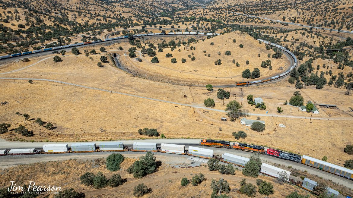 September 18th, 2024, BNSF 7115, 9128 and Ferromex 4050 lead an westbound train as they wait for an eastbound intermodal to clear Tehachapi Loop on the Union Pacific Mojave Subdivision at Keene, California.

I’m thankful that I had and brought my drone on this trip as access to trackside at the loop has been cut off! I was last here 7 years ago and then you could get to where you could shoot trackside, but now there’s viewing platform about ¾ mile away. Being able to fly the drone allowed me many spectacular views that I’ve never been able to get in the past.

According to Wikipedia: The Tehachapi Loop is a 3,779-foot-long (0.72 mi; 1.15 km) spiral, or helix, on the Union Pacific Railroad Mojave Subdivision through Tehachapi Pass, of the Tehachapi Mountains in Kern County, south-central California. The line connects Bakersfield and the San Joaquin Valley to Mojave in the Mojave Desert.

Rising at a steady two-percent grade, the track gains 77 feet (23 m) in elevation and makes a 1,210-foot-diameter (370 m) circle. Any train that is more than 3,800 feet (1,200 m) long—about 56 boxcars—passes over itself going around the loop. At the bottom of the loop, the track passes through Tunnel 9, the ninth tunnel built as the railroad was extended from Bakersfield.

The line averages about 36 freight trains each day. Passenger trains such as Amtrak's San Joaquin are banned from the loop, although the Coast Starlight can use it as a detour. Its frequent trains and scenic setting make the Tehachapi Loop popular with railfans. In 1998, it was named a National Historic Civil Engineering Landmark. It is also designated as California Historical Landmark #508.

One of the engineering feats of its day, the Loop was built by Southern Pacific Railroad to ease the grade over Tehachapi Pass. Construction began in 1874, and the line opened in 1876.

Tech Info: DJI Mavic 3 Classic Drone, RAW, 24mm, f/2.8, 1/2000, ISO 100.

#railroad #railroads #train, #trains #railway #railway #steamtrains #railtransport #railroadengines #picturesoftrains #picturesofrailways #besttrainphotograph #bestphoto #photographyoftrains #bestsoldpicture #JimPearsonPhotography #trainsfromtheair #trainsfromadrone #TehachapiLoop