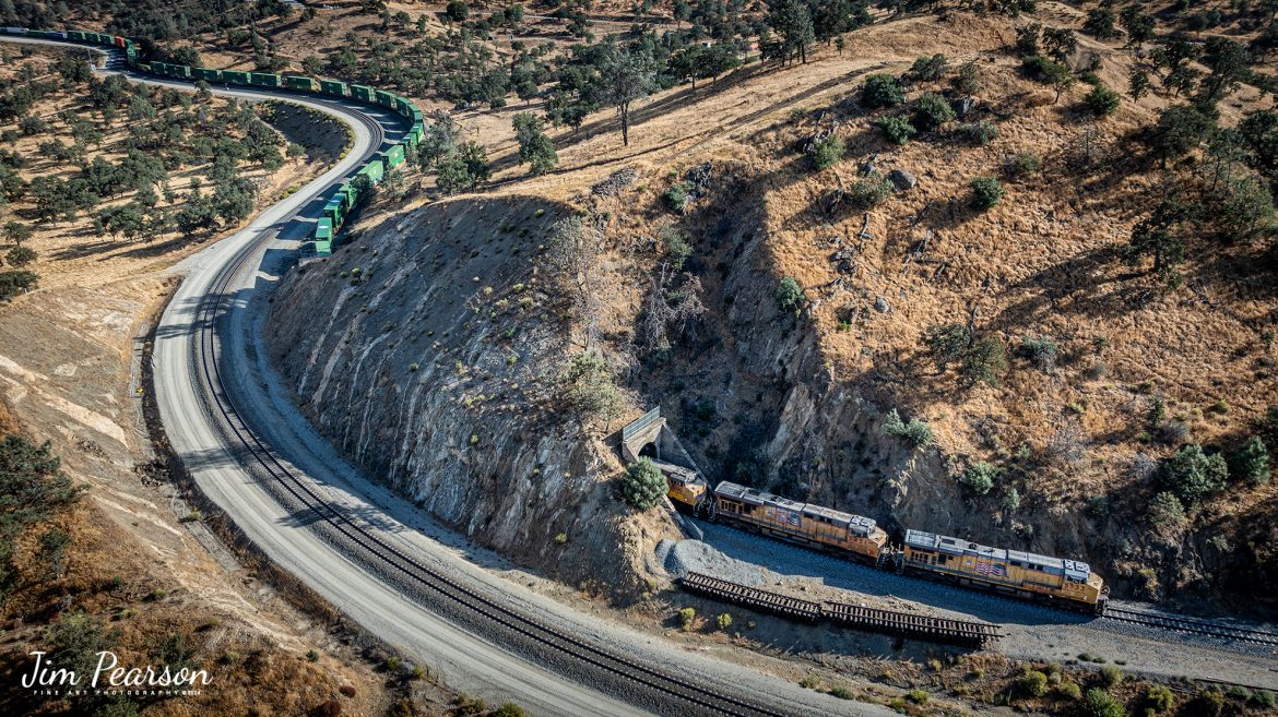 September 18th, 2024, Union Pacific 5533 leads an westbound intermodal train as they exit tunnel 10 at Tehachapi Loop on the Union Pacific Mojave Subdivision at Keene, California.

I’m thankful that I had and brought my drone on this trip as access to trackside at the loop has been cut off! I was last here 7 years ago and then you could get to where you could shoot trackside, but now there’s viewing platform about ¾ mile away. Being able to fly the drone allowed me many spectacular views that I’ve never been able to get in the past.

According to Wikipedia: The Tehachapi Loop is a 3,779-foot-long (0.72 mi; 1.15 km) spiral, or helix, on the Union Pacific Railroad Mojave Subdivision through Tehachapi Pass, of the Tehachapi Mountains in Kern County, south-central California. The line connects Bakersfield and the San Joaquin Valley to Mojave in the Mojave Desert.

Rising at a steady two-percent grade, the track gains 77 feet (23 m) in elevation and makes a 1,210-foot-diameter (370 m) circle. Any train that is more than 3,800 feet (1,200 m) long—about 56 boxcars—passes over itself going around the loop. At the bottom of the loop, the track passes through Tunnel 9, the ninth tunnel built as the railroad was extended from Bakersfield.

The line averages about 36 freight trains each day. Passenger trains such as Amtrak's San Joaquin are banned from the loop, although the Coast Starlight can use it as a detour. Its frequent trains and scenic setting make the Tehachapi Loop popular with railfans. In 1998, it was named a National Historic Civil Engineering Landmark. It is also designated as California Historical Landmark #508.

One of the engineering feats of its day, the Loop was built by Southern Pacific Railroad to ease the grade over Tehachapi Pass. Construction began in 1874, and the line opened in 1876.

Tech Info: DJI Mavic 3 Classic Drone, RAW, 24mm, f/2.8, 1/800, ISO 100.

#railroad #railroads #train, #trains #railway #railway #steamtrains #railtransport #railroadengines #picturesoftrains #picturesofrailways #besttrainphotograph #bestphoto #photographyoftrains #bestsoldpicture #JimPearsonPhotography #trainsfromtheair #trainsfromadrone #TehachapiLoop