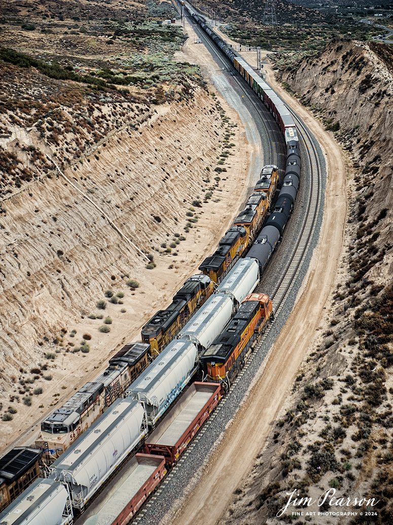 September 20th, 2024, Today I had the good fortune of catching a three way meet between a Union Pacific Freight, a BNSF freight and a BNSF rock train at the Summit of Cajon Pass in southern California and the BNSF Cajon Subdivision as Hwy 138 and the tracks head into stormy weather.

According to Wikipedia: Cajon Pass is a mountain pass between the San Bernardino Mountains to the east and the San Gabriel Mountains to the west in Southern California. Created by the movements of the San Andreas Fault, it has an elevation of 3,777 ft (1,151 m). Located in the Mojave Desert, the pass is an important link from the Greater San Bernardino Area to the Victor Valley, and northeast to Las Vegas. The Cajon Pass area is on the Pacific Crest Trail.

Cajon Pass is at the head of Horsethief Canyon, traversed by California State Route 138 (SR 138) and railroad tracks owned by BNSF Railway and Union Pacific Railroad. Improvements in 1972 reduced the railroad's maximum elevation from about 3,829 to 3,777 feet while reducing curvature. Interstate 15 does not traverse Cajon Pass, but rather the nearby Cajon Summit. The entire area, Cajon Pass and Cajon Summit, is often referred to as Cajon Pass, but a distinction is made between Cajon Pass and Cajon Summit.

The California Southern Railroad, a subsidiary of the Atchison, Topeka and Santa Fe Railway, was the first railroad through Cajon Pass. The line through the pass was built in the early 1880s to connect the present-day cities of Barstow and San Diego. Today the Union Pacific Railroad and BNSF Railway (the successor to the Santa Fe) use the pass to reach Los Angeles and San Bernardino as part of the Southern Transcon. Due to the many trains, scenery and easy access, it is a popular location for railfans, and many photographs of trains on Cajon Pass appear in books and magazines.

The Union Pacific Railroad owns one track through the pass, on the previous Southern Pacific Railroad Palmdale cutoff, opened in 1967. The BNSF Railway owns two tracks and began to operate a third main track in the summer of 2008. The railroads share track rights through the pass ever since the Union Pacific gained track rights on the Santa Fe portion negotiated under the original Los Angeles and Salt Lake Railroad. 

Tech Info: DJI Mavic 3 Classic Drone, RAW, 24mm, f/2.8, 1/1250, ISO 100.

#railroad #railroads #train, #trains #railway #railway #steamtrains #railtransport #railroadengines #picturesoftrains #picturesofrailways #besttrainphotograph #bestphoto #photographyoftrains #bestsoldpicture #JimPearsonPhotography #trainsfromtheair #trainsfromadrone #CajonPass