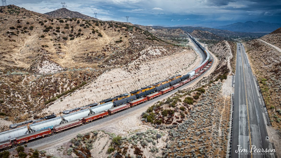 September 20th, 2024, Today I had the good fortune of catching a three way meet between a Union Pacific Freight, a BNSF freight and a BNSF rock train at the Summit of Cajon Pass in southern California and the BNSF Cajon Subdivision as Hwy 138 and the tracks head into stormy weather.

According to Wikipedia: Cajon Pass is a mountain pass between the San Bernardino Mountains to the east and the San Gabriel Mountains to the west in Southern California. Created by the movements of the San Andreas Fault, it has an elevation of 3,777 ft (1,151 m). Located in the Mojave Desert, the pass is an important link from the Greater San Bernardino Area to the Victor Valley, and northeast to Las Vegas. The Cajon Pass area is on the Pacific Crest Trail.

Cajon Pass is at the head of Horsethief Canyon, traversed by California State Route 138 (SR 138) and railroad tracks owned by BNSF Railway and Union Pacific Railroad. Improvements in 1972 reduced the railroad's maximum elevation from about 3,829 to 3,777 feet while reducing curvature. Interstate 15 does not traverse Cajon Pass, but rather the nearby Cajon Summit. The entire area, Cajon Pass and Cajon Summit, is often referred to as Cajon Pass, but a distinction is made between Cajon Pass and Cajon Summit.

The California Southern Railroad, a subsidiary of the Atchison, Topeka and Santa Fe Railway, was the first railroad through Cajon Pass. The line through the pass was built in the early 1880s to connect the present-day cities of Barstow and San Diego. Today the Union Pacific Railroad and BNSF Railway (the successor to the Santa Fe) use the pass to reach Los Angeles and San Bernardino as part of the Southern Transcon. Due to the many trains, scenery and easy access, it is a popular location for railfans, and many photographs of trains on Cajon Pass appear in books and magazines.

The Union Pacific Railroad owns one track through the pass, on the previous Southern Pacific Railroad Palmdale cutoff, opened in 1967. The BNSF Railway owns two tracks and began to operate a third main track in the summer of 2008. The railroads share track rights through the pass ever since the Union Pacific gained track rights on the Santa Fe portion negotiated under the original Los Angeles and Salt Lake Railroad. 

Tech Info: DJI Mavic 3 Classic Drone, RAW, 24mm, f/2.8, 1/1250, ISO 100.

#railroad #railroads #train, #trains #railway #railway #steamtrains #railtransport #railroadengines #picturesoftrains #picturesofrailways #besttrainphotograph #bestphoto #photographyoftrains #bestsoldpicture #JimPearsonPhotography #trainsfromtheair #trainsfromadrone #CajonPass