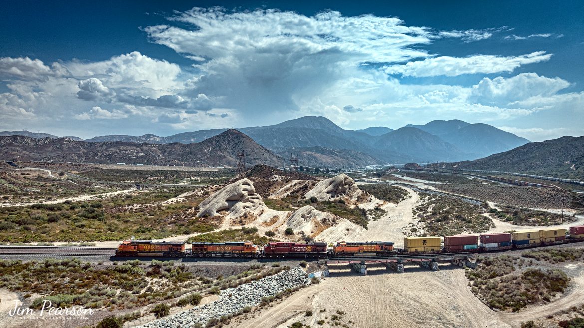 BNSF 4037, 4549, CP 8125 and BNSF 5733 lead an eastbound intermodal at Phelan, CA, as they cross the dry wash east of Hwy 138 in the Cajon Pass on September 20th, 2024, on the BNSF Cajon Subdivision.   

According to Wikipedia: Cajon Pass is a mountain pass between the San Bernardino Mountains to the east and the San Gabriel Mountains to the west in Southern California. Created by the movements of the San Andreas Fault, it has an elevation of 3,777 ft (1,151 m). Located in the Mojave Desert, the pass is an important link from the Greater San Bernardino Area to the Victor Valley, and northeast to Las Vegas. The Cajon Pass area is on the Pacific Crest Trail.

Cajon Pass is at the head of Horsethief Canyon, traversed by California State Route 138 (SR 138) and railroad tracks owned by BNSF Railway and Union Pacific Railroad. Improvements in 1972 reduced the railroad's maximum elevation from about 3,829 to 3,777 feet while reducing curvature. Interstate 15 does not traverse Cajon Pass, but rather the nearby Cajon Summit. The entire area, Cajon Pass and Cajon Summit, is often referred to as Cajon Pass, but a distinction is made between Cajon Pass and Cajon Summit.

The California Southern Railroad, a subsidiary of the Atchison, Topeka and Santa Fe Railway, was the first railroad through Cajon Pass. The line through the pass was built in the early 1880s to connect the present-day cities of Barstow and San Diego. Today the Union Pacific Railroad and BNSF Railway (the successor to the Santa Fe) use the pass to reach Los Angeles and San Bernardino as part of the Southern Transcon. Due to the many trains, scenery and easy access, it is a popular location for railfans, and many photographs of trains on Cajon Pass appear in books and magazines.

The Union Pacific Railroad owns one track through the pass, on the previous Southern Pacific Railroad Palmdale cutoff, opened in 1967. The BNSF Railway owns two tracks and began to operate a third main track in the summer of 2008. The railroads share track rights through the pass ever since the Union Pacific gained track rights on the Santa Fe portion negotiated under the original Los Angeles and Salt Lake Railroad. 

Tech Info: DJI Mavic 3 Classic Drone, RAW, 24mm, f/2.8, 1/4000, ISO 140.

#railroad #railroads #train, #trains #railway #railway #steamtrains #railtransport #railroadengines #picturesoftrains #picturesofrailways #besttrainphotograph #bestphoto #photographyoftrains #bestsoldpicture #JimPearsonPhotography #trainsfromtheair #trainsfromadrone #CajonPass