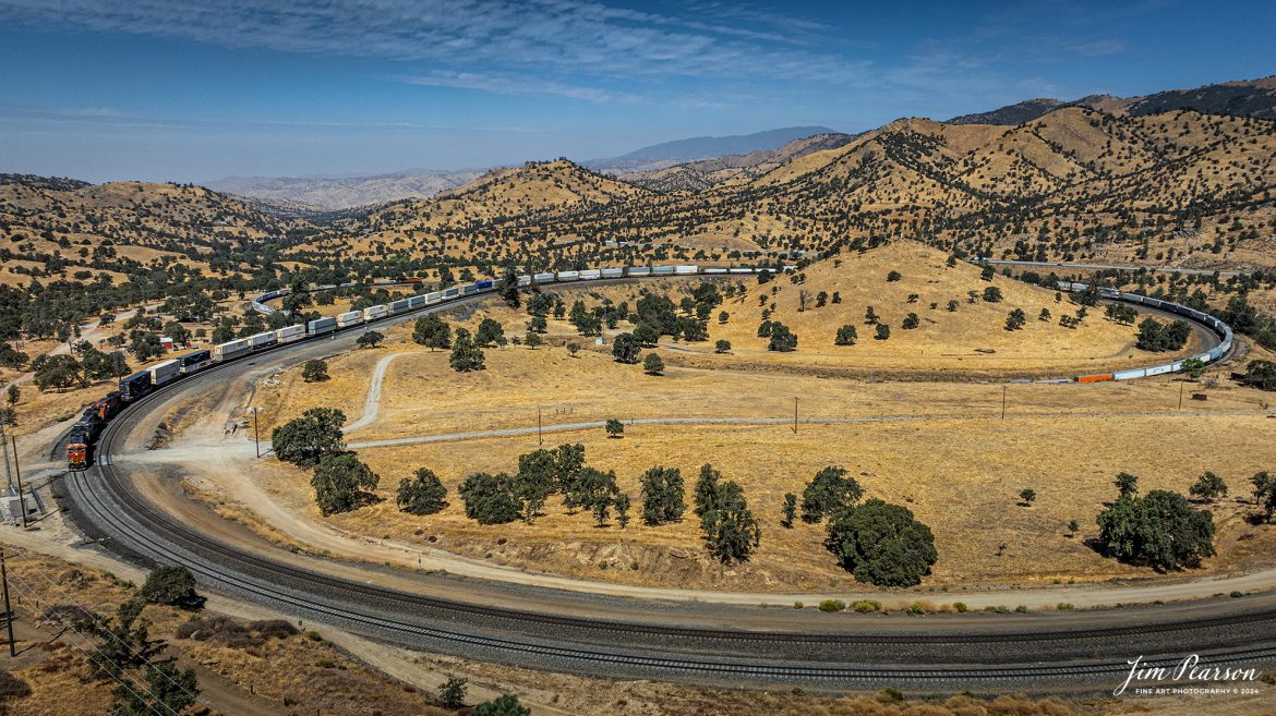 September 18th, 2024, BNSF 8057, NS 8068 and BNSF 5177 lead an eastbound train through the famous Tehachapi Loop on the Union Pacific Mojave Subdivision at Keene, California.

According to Wikipedia: The Tehachapi Loop is a 3,779-foot-long (0.72 mi; 1.15 km) spiral, or helix, on the Union Pacific Railroad Mojave Subdivision through Tehachapi Pass, of the Tehachapi Mountains in Kern County, south-central California. The line connects Bakersfield and the San Joaquin Valley to Mojave in the Mojave Desert.

Rising at a steady two-percent grade, the track gains 77 feet (23 m) in elevation and makes a 1,210-foot-diameter (370 m) circle. Any train that is more than 3,800 feet (1,200 m) long—about 56 boxcars—passes over itself going around the loop. At the bottom of the loop, the track passes through Tunnel 9, the ninth tunnel built as the railroad was extended from Bakersfield.

The line averages about 36 freight trains each day. Passenger trains such as Amtrak's San Joaquin are banned from the loop, although the Coast Starlight can use it as a detour. Its frequent trains and scenic setting make the Tehachapi Loop popular with railfans. In 1998, it was named a National Historic Civil Engineering Landmark. It is also designated as California Historical Landmark #508.

One of the engineering feats of its day, the Loop was built by Southern Pacific Railroad to ease the grade over Tehachapi Pass. Construction began in 1874, and the line opened in 1876.

Tech Info: DJI Mavic 3 Classic Drone, RAW, 24mm, f/2.8, 1/2500, ISO 100.

#railroad #railroads #train, #trains #railway #railway #steamtrains #railtransport #railroadengines #picturesoftrains #picturesofrailways #besttrainphotograph #bestphoto #photographyoftrains #bestsoldpicture #JimPearsonPhotography #trainsfromtheair #trainsfromadrone #TehachapiLoop