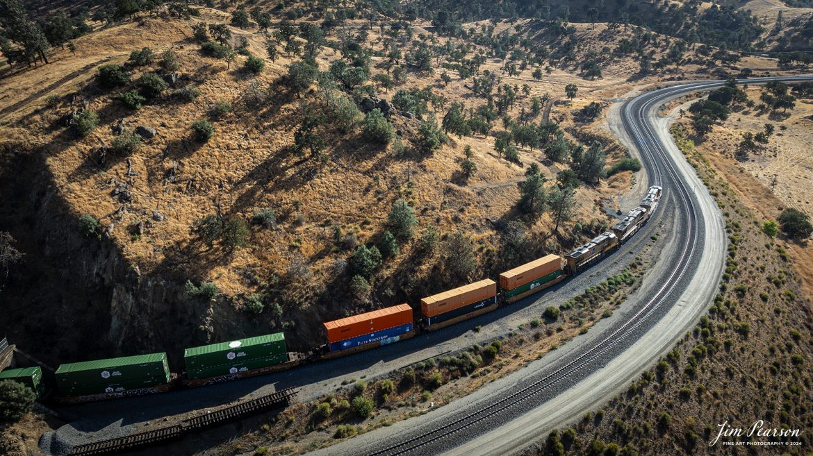 September 18th, 2024, Union Pacific 5533 leads an southbound intermodal train as they exit tunnel 10 and head into Tehachapi Loop on the Union Pacific Mojave Subdivision at Woodford, California.

According to Wikipedia: The Tehachapi Loop is a 3,779-foot-long (0.72 mi; 1.15 km) spiral, or helix, on the Union Pacific Railroad Mojave Subdivision through Tehachapi Pass, of the Tehachapi Mountains in Kern County, south-central California. The line connects Bakersfield and the San Joaquin Valley to Mojave in the Mojave Desert.

Rising at a steady two-percent grade, the track gains 77 feet (23 m) in elevation and makes a 1,210-foot-diameter (370 m) circle. Any train that is more than 3,800 feet (1,200 m) long—about 56 boxcars—passes over itself going around the loop. At the bottom of the loop, the track passes through Tunnel 9, the ninth tunnel built as the railroad was extended from Bakersfield.

The line averages about 36 freight trains each day. Passenger trains such as Amtrak's San Joaquin are banned from the loop, although the Coast Starlight can use it as a detour. Its frequent trains and scenic setting make the Tehachapi Loop popular with railfans. In 1998, it was named a National Historic Civil Engineering Landmark. It is also designated as California Historical Landmark #508.

One of the engineering feats of its day, the Loop was built by Southern Pacific Railroad to ease the grade over Tehachapi Pass. Construction began in 1874, and the line opened in 1876.

Tech Info: DJI Mavic 3 Classic Drone, RAW, 24mm, f/2.8, 1/800, ISO 110.

#railroad #railroads #train, #trains #railway #railway #steamtrains #railtransport #railroadengines #picturesoftrains #picturesofrailways #besttrainphotograph #bestphoto #photographyoftrains #bestsoldpicture #JimPearsonPhotography #trainsfromtheair #trainsfromadrone #TehachapiLoop