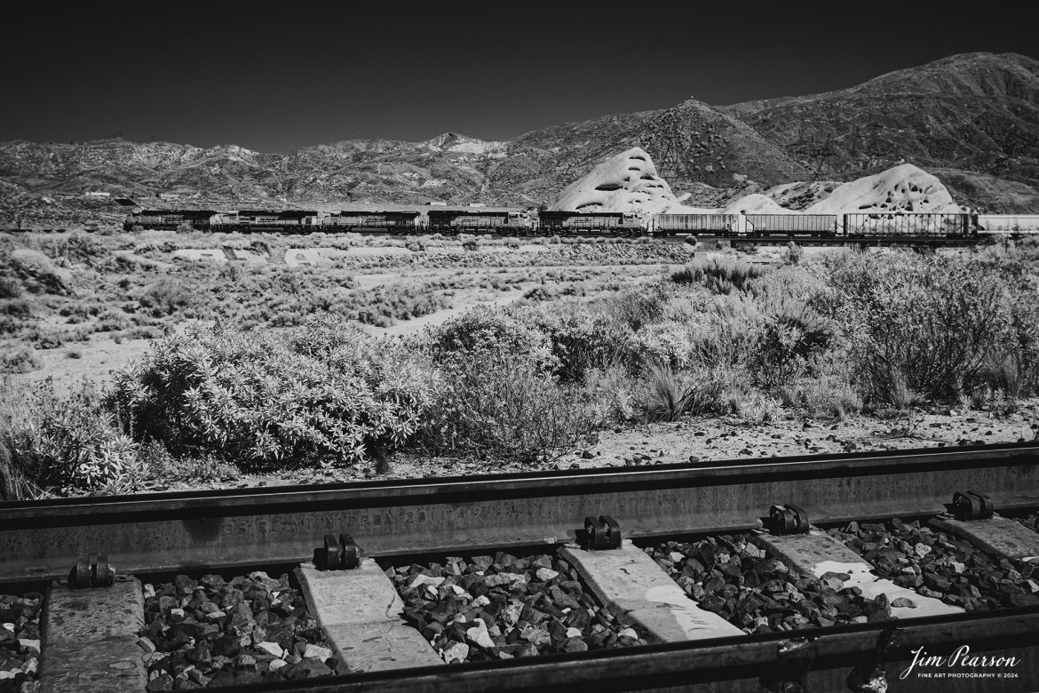 In this week’s Saturday Infrared photo, we catch a loaded BNSF freight as it starts the climb up the grade in the Cajon Pass from Cajon, California on September 24th, 2024.

According to Wikipedia: Cajon Pass is a mountain pass between the San Bernardino Mountains to the east and the San Gabriel Mountains to the west in Southern California. Created by the movements of the San Andreas Fault, it has an elevation of 3,777 ft (1,151 m). Located in the Mojave Desert, the pass is an important link from the Greater San Bernardino Area to the Victor Valley, and northeast to Las Vegas. The Cajon Pass area is on the Pacific Crest Trail.

Cajon Pass is at the head of Horsethief Canyon, traversed by California State Route 138 (SR 138) and railroad tracks owned by BNSF Railway and Union Pacific Railroad. Improvements in 1972 reduced the railroad's maximum elevation from about 3,829 to 3,777 feet while reducing curvature. Interstate 15 does not traverse Cajon Pass, but rather the nearby Cajon Summit. The entire area, Cajon Pass and Cajon Summit, is often referred to as Cajon Pass, but a distinction is made between Cajon Pass and Cajon Summit.

The California Southern Railroad, a subsidiary of the Atchison, Topeka and Santa Fe Railway, was the first railroad through Cajon Pass. The line through the pass was built in the early 1880s to connect the present-day cities of Barstow and San Diego. Today the Union Pacific Railroad and BNSF Railway (the successor to the Santa Fe) use the pass to reach Los Angeles and San Bernardino as part of the Southern Transcon. Due to the many trains, scenery and easy access, it is a popular location for railfans, and many photographs of trains on Cajon Pass appear in books and magazines.

The Union Pacific Railroad owns one track through the pass, on the previous Southern Pacific Railroad Palmdale cutoff, opened in 1967. The BNSF Railway owns two tracks and began to operate a third main track in the summer of 2008. The railroads share track rights through the pass ever since the Union Pacific gained track rights on the Santa Fe portion negotiated under the original Los Angeles and Salt Lake Railroad. 

Tech Info: Fuji XT-1, RAW, Converted to 720nm B&W IR, Nikon 24-70 @28mm, f/4.5, 1/500, ISO 200.

#trainphotography #railroadphotography #trains #railways #jimpearsonphotography #infraredtrainphotography #infraredphotography #trainphotographer #railroadphotographer #infaredtrainphotography #trending