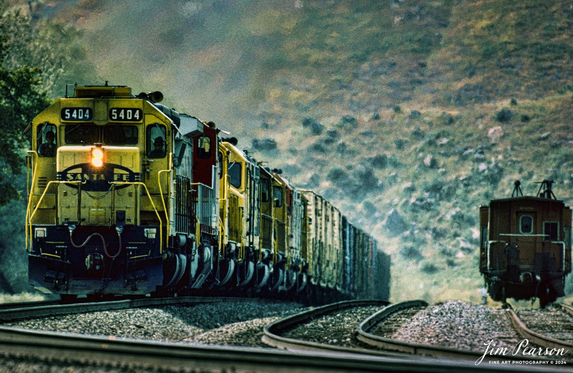 Santa Fe 5404 leads 8 locomotives as they climb upgrade through the Tehachapi Pass in central California in during early 1995 passing a caboose parked in a house track. Unfortunately I can't remember off the top of my head where this spot is at or what it's called. Anyone know?

According to Wikipedia: Tehachapi Pass (Kawaiisu: Tihachipia, meaning "hard climb") is a mountain pass crossing the Tehachapi Mountains in Kern County, California. Traditionally, the pass marks the northeast end of the Tehachapi’s and the south end of the Sierra Nevada range.

The route is a principal connector between the San Joaquin Valley and the Mojave Desert. The Native American Kitanemuk people used the pass as a trade route before the American settlement of the region in the 19th century. The main line of the former Southern Pacific Railroad opened though the pass in 1876;[4] the tracks are now owned by the Union Pacific Railroad and shared with BNSF Railway as the Mojave Subdivision. U.S. Route 466 was built in the 1930s, and the road is now State Route 58. The Pass is also the route of the planned California High-Speed Rail line.

Tech Notes: Nikon F3 Film Camera, Nikon 300mm, f/stop and shutter speed not recorded

#railroad #railroads #train #trains #bestphoto #railroadengines #picturesoftrains #picturesofrailway #bestphotograph #photographyoftrains #trainphotography #JimPearsonPhotography