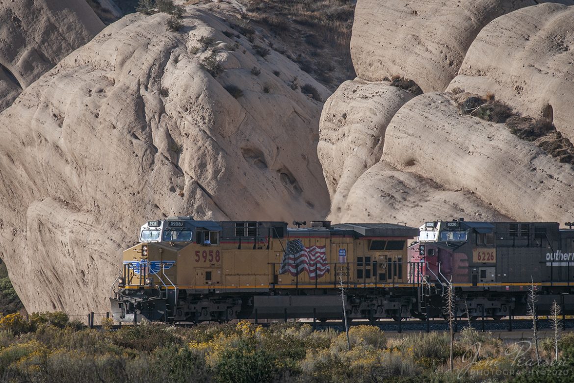 October 20, 2008 - The late evening light rakes across the base of Mormon Rocks as UP 5958 and 6266 (ex-Southern Pacific Unit) lead a west bound freight down the Cajon Pass in southern California. 

Local legend has it that the rocks were named for the Mormon pioneers who camped here after their descent from the pass ridgeline. Another legend calls the rocks the 'Chanting Rocks,' as when the wind would blow across the portholes in the rocks it was said the sound made was similar to a low chanting or singing.

Nikon D300, Nikon 70-300 @ 300mm, 1/640, f/8, ISO 200, RAW.