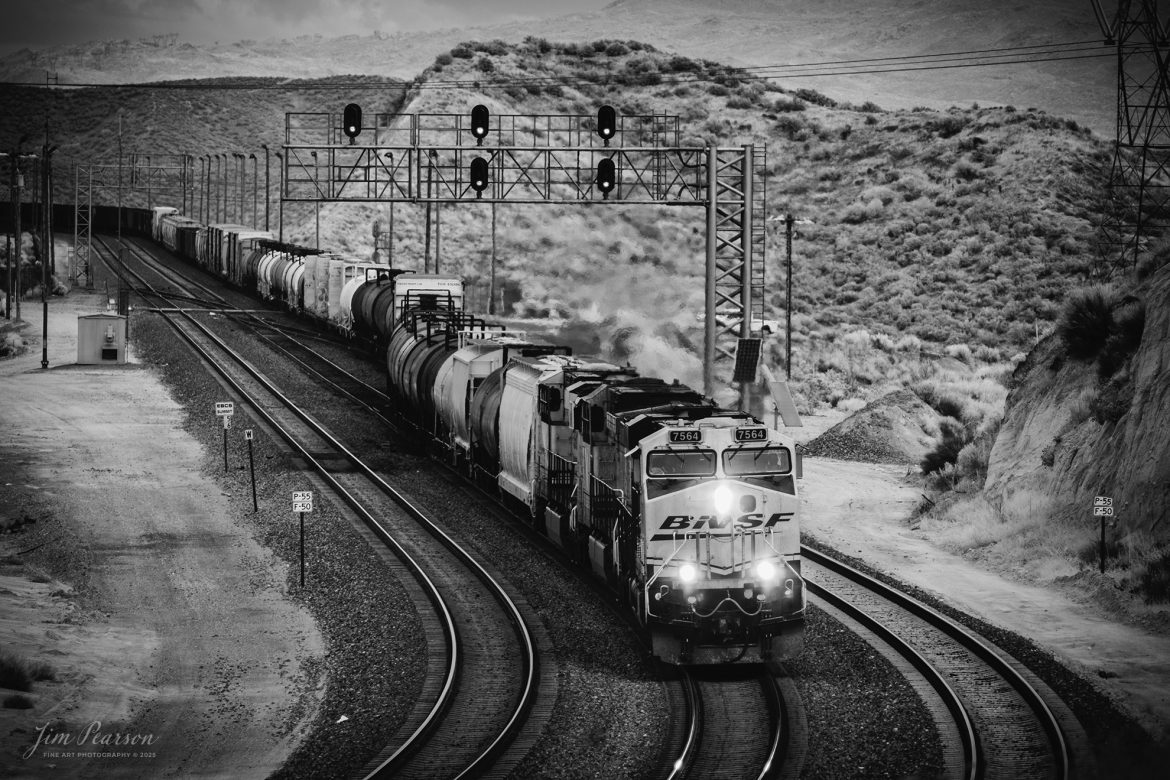 BNSF 7564 leads an downhill freight as it passes through a crossover at Summit in the Cajon Pass at Cajon, California on the BNSF Cajon Subdivision, on September 20th, 2024. 

According to Wikipedia: Cajon Pass is a mountain pass between the San Bernardino Mountains to the east and the San Gabriel Mountains to the west in Southern California. Created by the movements of the San Andreas Fault, it has an elevation of 3,777 ft (1,151 m). Located in the Mojave Desert, the pass is an important link from the Greater San Bernardino Area to the Victor Valley, and northeast to Las Vegas. The Cajon Pass area is on the Pacific Crest Trail.

Cajon Pass is at the head of Horsethief Canyon, traversed by California State Route 138 (SR 138) and railroad tracks owned by BNSF Railway and Union Pacific Railroad. Improvements in 1972 reduced the railroad's maximum elevation from about 3,829 to 3,777 feet while reducing curvature. Interstate 15 does not traverse Cajon Pass, but rather the nearby Cajon Summit. The entire area, Cajon Pass and Cajon Summit, is often referred to as Cajon Pass, but a distinction is made between Cajon Pass and Cajon Summit.

The California Southern Railroad, a subsidiary of the Atchison, Topeka and Santa Fe Railway, was the first railroad through Cajon Pass. The line through the pass was built in the early 1880s to connect the present-day cities of Barstow and San Diego. Today the Union Pacific Railroad and BNSF Railway (the successor to the Santa Fe) use the pass to reach Los Angeles and San Bernardino as part of the Southern Transcon. Due to the many trains, scenery and easy access, it is a popular location for railfans, and many photographs of trains on Cajon Pass appear in books and magazines.

The Union Pacific Railroad owns one track through the pass, on the previous Southern Pacific Railroad Palmdale cutoff, opened in 1967. The BNSF Railway owns two tracks and began to operate a third main track in the summer of 2008. The railroads share track rights through the pass ever since the Union Pacific gained track rights on the Santa Fe portion negotiated under the original Los Angeles and Salt Lake Railroad. 

Tech Info: Fuji XT-1, RAW, Converted to 720nm B&W IR, Nikon 70-300 @200mm, f/5.6, 1/180, ISO 200.

#trainphotography #railroadphotography #trains #railways #jimpearsonphotography #infraredtrainphotography #infraredphotography #trainphotographer #railroadphotographer #infaredtrainphotography #trending