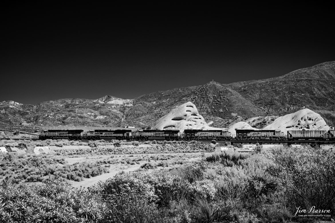 In this week’s Saturday Infrared photo, we catch a loaded BNSF freight as works the grade in the Cajon Pass from Cajon, California on September 24th, 2024.

According to Wikipedia: Cajon Pass is a mountain pass between the San Bernardino Mountains to the east and the San Gabriel Mountains to the west in Southern California. Created by the movements of the San Andreas Fault, it has an elevation of 3,777 ft (1,151 m). Located in the Mojave Desert, the pass is an important link from the Greater San Bernardino Area to the Victor Valley, and northeast to Las Vegas. The Cajon Pass area is on the Pacific Crest Trail.

Cajon Pass is at the head of Horsethief Canyon, traversed by California State Route 138 (SR 138) and railroad tracks owned by BNSF Railway and Union Pacific Railroad. Improvements in 1972 reduced the railroad's maximum elevation from about 3,829 to 3,777 feet while reducing curvature. Interstate 15 does not traverse Cajon Pass, but rather the nearby Cajon Summit. The entire area, Cajon Pass and Cajon Summit, is often referred to as Cajon Pass, but a distinction is made between Cajon Pass and Cajon Summit.

The California Southern Railroad, a subsidiary of the Atchison, Topeka and Santa Fe Railway, was the first railroad through Cajon Pass. The line through the pass was built in the early 1880s to connect the present-day cities of Barstow and San Diego. Today the Union Pacific Railroad and BNSF Railway (the successor to the Santa Fe) use the pass to reach Los Angeles and San Bernardino as part of the Southern Transcon. Due to the many trains, scenery and easy access, it is a popular location for railfans, and many photographs of trains on Cajon Pass appear in books and magazines.

The Union Pacific Railroad owns one track through the pass, on the previous Southern Pacific Railroad Palmdale cutoff, opened in 1967. The BNSF Railway owns two tracks and began to operate a third main track in the summer of 2008. The railroads share track rights through the pass ever since the Union Pacific gained track rights on the Santa Fe portion negotiated under the original Los Angeles and Salt Lake Railroad. 

Tech Info: Fuji XT-1, RAW, Converted to 720nm B&W IR, Nikon 24-70 @39mm, f/4.5, 1/500, ISO 200.

#trainphotography #railroadphotography #trains #railways #jimpearsonphotography #infraredtrainphotography #infraredphotography #trainphotographer #railroadphotographer #infaredtrainphotography #trending